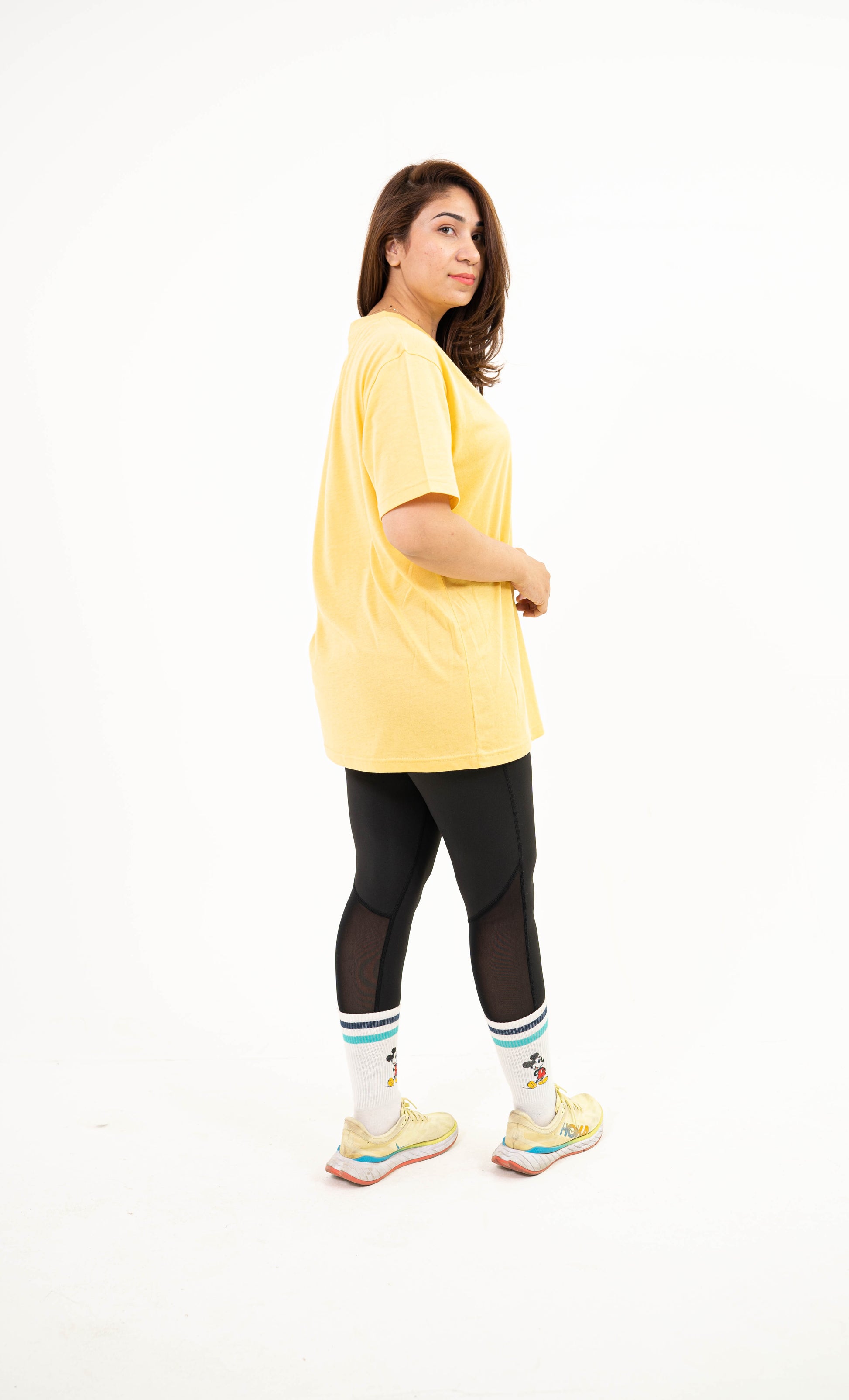 A woman modeling an oversized pale yellow t-shirt, black leggings, white socks with teal stripes, and yellow sneakers, photographed against a seamless white studio background.