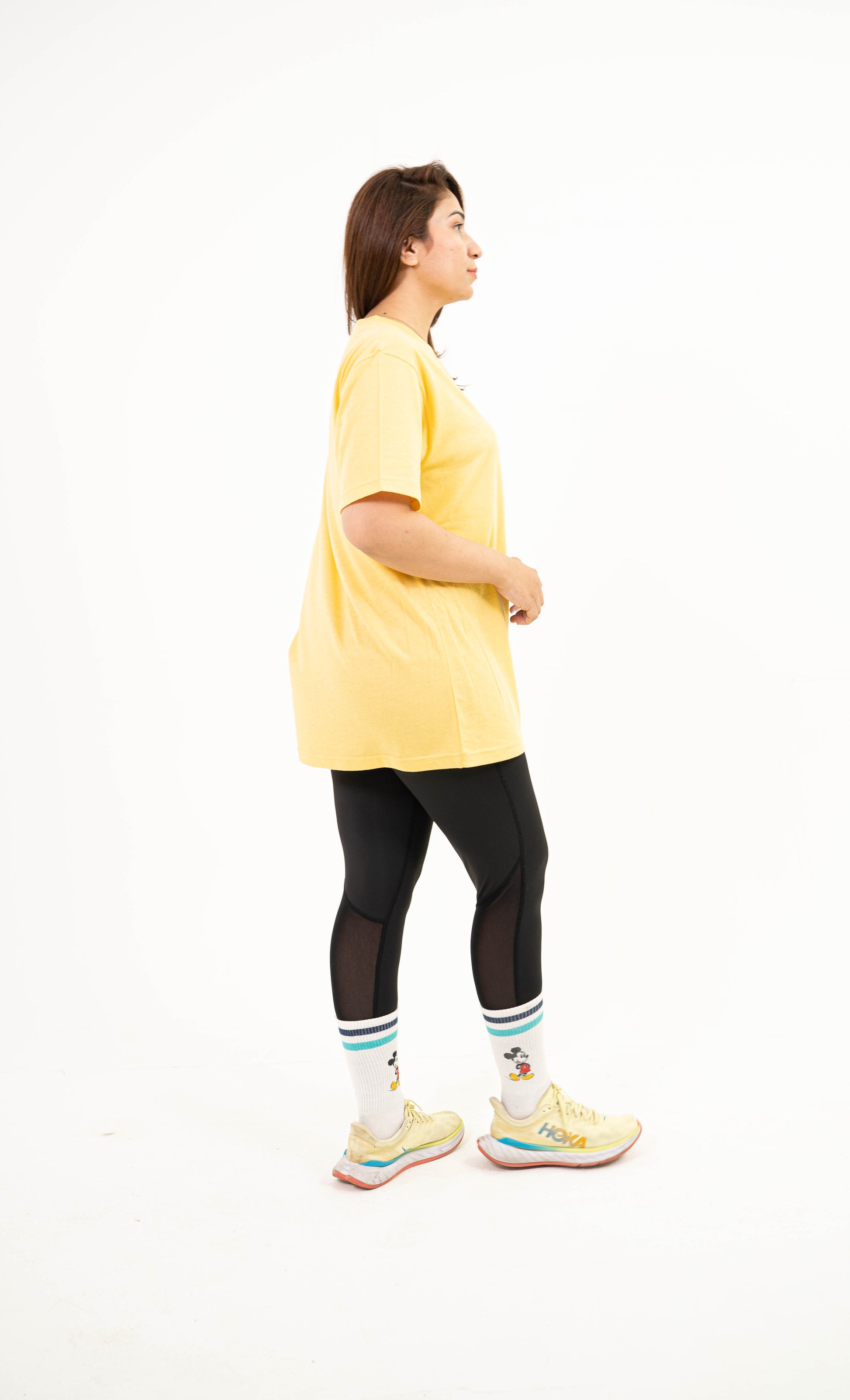 A woman shown in side profile wearing a loose yellow short-sleeve top, black leggings, white socks with green and blue stripes, and light yellow running shoes against a plain white studio background.