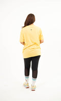 A person viewed from behind wearing a bright yellow oversized short-sleeve T-shirt, black leggings, white socks, and light-colored sneakers, standing against a plain white studio background.