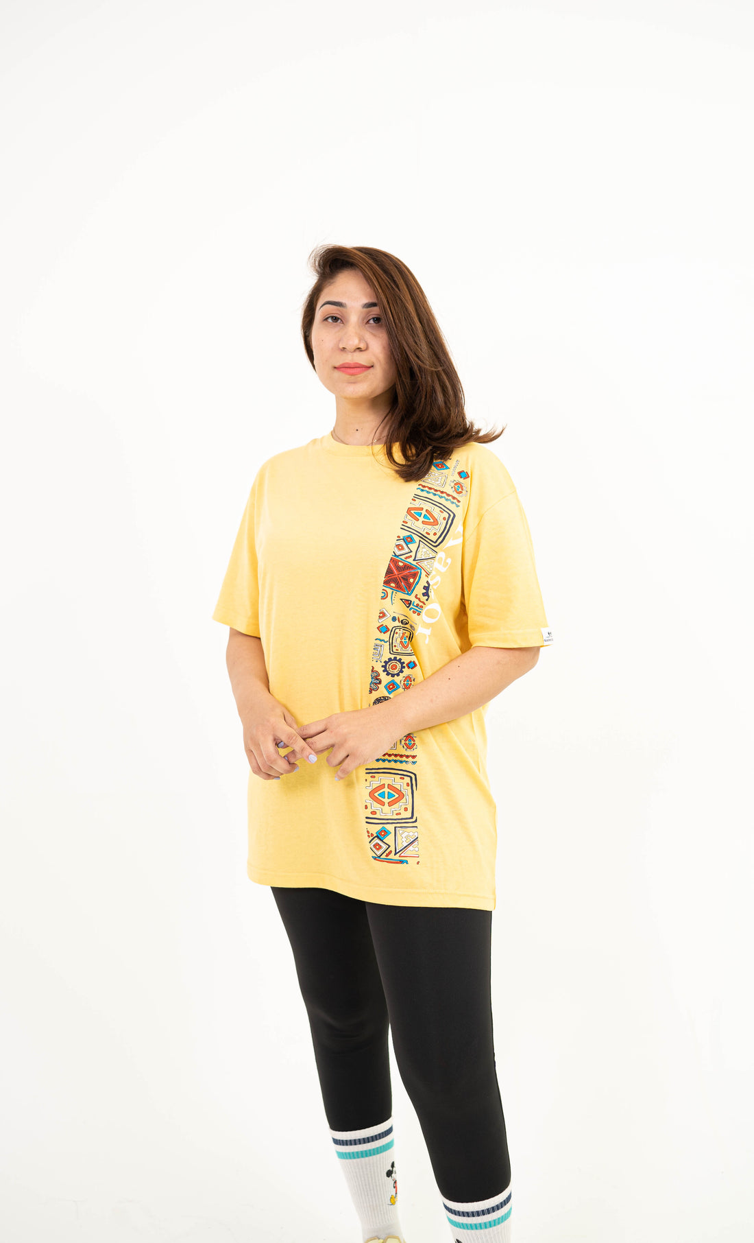 A woman modeling a yellow oversized t-shirt with a vertical multicolor graphic print down the right side, paired with black leggings, white socks, and yellow sneakers, against a plain white background.