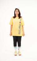 A woman modeling a yellow oversized t-shirt with a vertical multicolor graphic print down the right side, paired with black leggings, white socks, and yellow sneakers, against a plain white background.