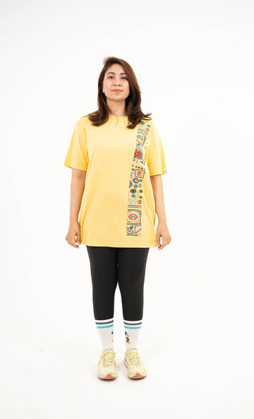 A woman modeling a yellow oversized t-shirt with a vertical multicolor graphic print down the right side, paired with black leggings, white socks, and yellow sneakers, against a plain white background.