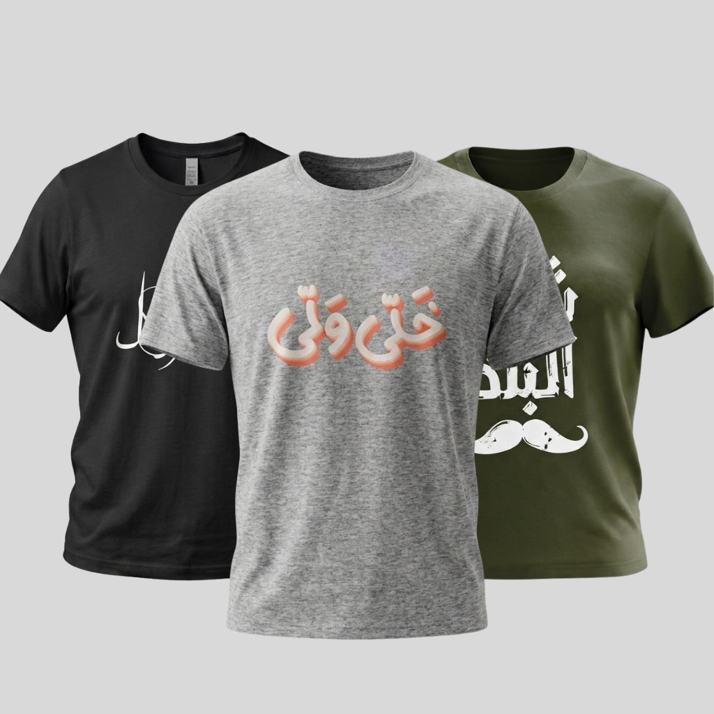 Three short-sleeve crew-neck t-shirts displayed together on a light gray background: a black shirt on the left with white Arabic calligraphy, a heather gray shirt in the center with peach Arabic text across the chest, and an olive green shirt on the right with white Arabic text and a white mustache graphic.