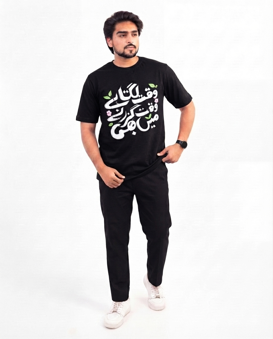 A man modeling a black short-sleeve t-shirt with white and green Arabic calligraphy on the front, paired with black pants and white sneakers, against a plain white studio background.