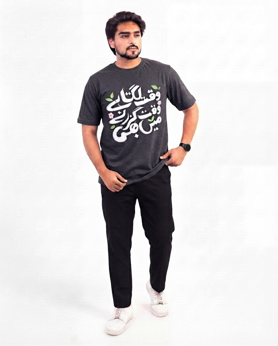 A man modeling an olive green graphic t-shirt with black Arabic calligraphy and small green and pink floral accents, paired with black pants and white sneakers, against a plain white background.