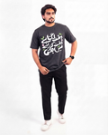 A man modeling a dark gray graphic t-shirt with white and green Arabic calligraphy, paired with black pants and white sneakers, against a plain white background.