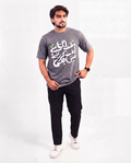 A man modeling a heather gray t-shirt with white and green Arabic calligraphy on the front, paired with black pants and white sneakers, against a plain white studio background.
