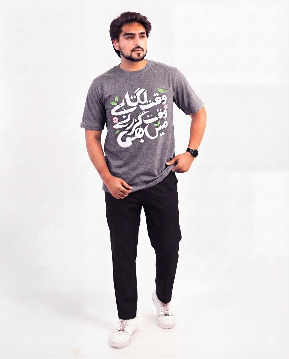 A man modeling a heather gray t-shirt with white and green Arabic calligraphy on the front, paired with black pants and white sneakers, against a plain white studio background.