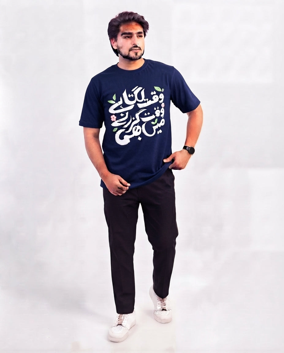 A man modeling a navy blue t-shirt with white Arabic calligraphy and small green and pink floral accents, paired with black pants and white sneakers, against a light gray studio background.