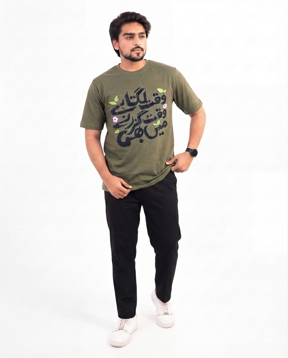 A man modeling an olive green graphic t-shirt with black Arabic calligraphy and small green and pink floral accents, paired with black pants and white sneakers, against a plain white background.